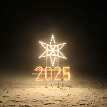 Ein beleuchtetes Sternschild mit der Zahl 2024 steht auf einer sandigen Oberfläche. Der Stern besteht aus leuchtenden Lichtern. Der Hintergrund ist dunkel.