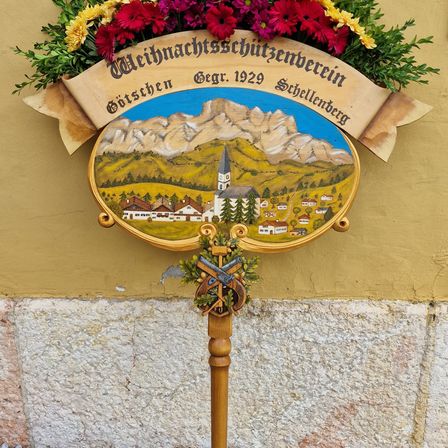 Ein bemaltes Schild mit Blumendekoration zeigt eine Szene von Bergen und einem Dorf, datiert 1929. Es ist an einem Holzpfahl montiert.