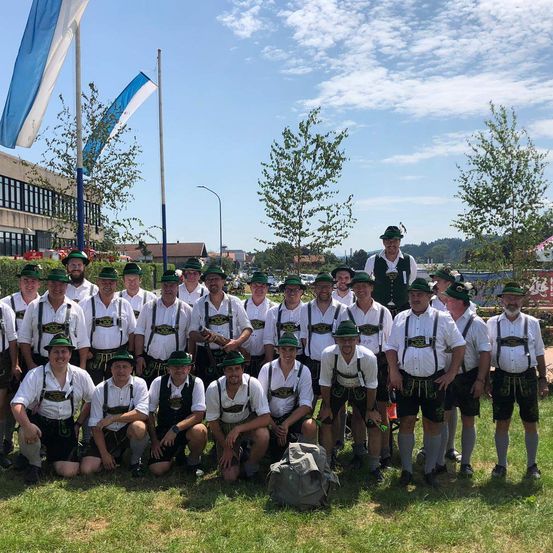 Eine Gruppe von Männern in traditioneller deutscher Kleidung posiert für ein Foto auf einer Wiese, mit Fahnen und Bäumen im Hintergrund.