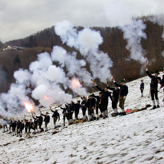 Eine Gruppe von Personen in traditioneller Kleidung, die Gewehre halten, steht auf einem verschneiten Hügel. Sie feuern mit den Gewehren in die Luft und erzeugen Rauch. Im Hintergrund ist ein Wald mit einem Haus in der Ferne zu sehen.