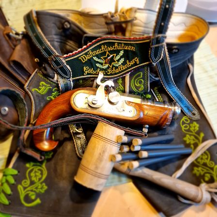 Eine Ledertasche mit traditioneller Stickerei enthält eine Vintage-Pistole mit Holzgriff, mehrere Kugeln und einen kleinen Hammer auf einem Holztisch.