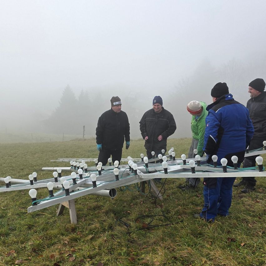 Mehrere Männer stehen in einem nebligen Feld, einige tragen Mützen und Handschuhe. Sie arbeiten an einer Struktur mit vielen angebrachten Glühbirnen.