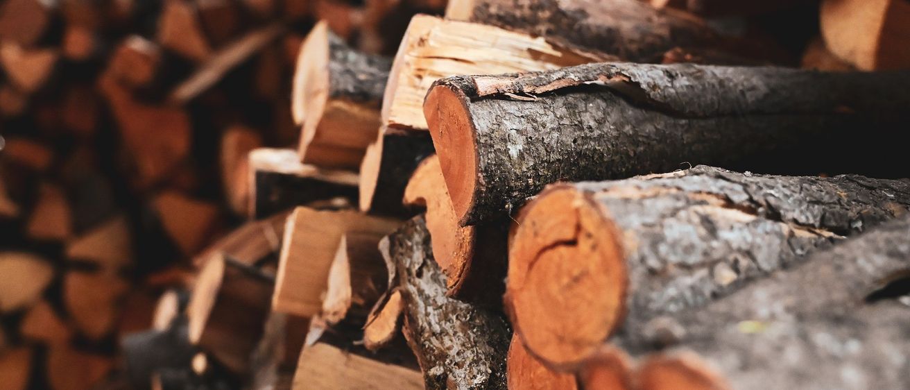 Ein Stapel Brennholz in verschiedenen Größen und Farben, einige mit Rinde, andere geschnitten. Die Holzscheite sind in einem Haufen angeordnet, beleuchtet von einem warmen, orangefarbenen Licht.