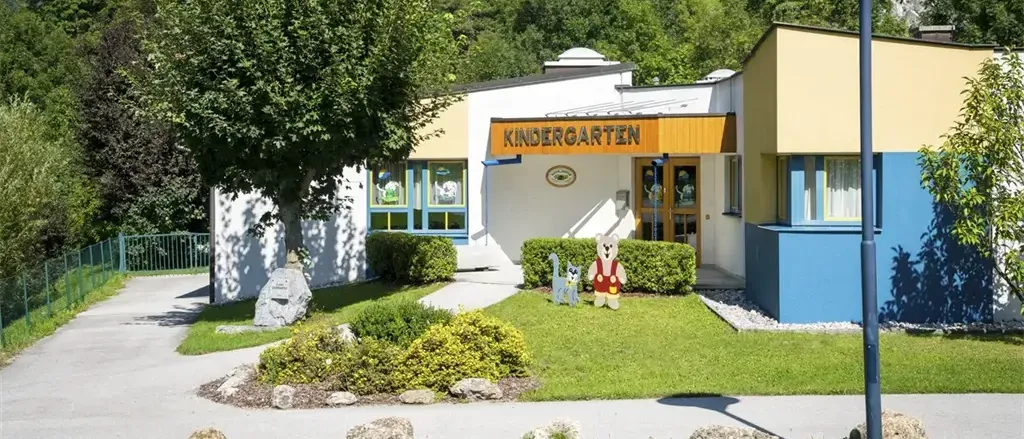 Ein Kindergarten mit einem gelben Schild, umgeben von einem Garten mit zwei Cartoon-Figuren davor. Bäume und Büsche zieren das Gelände, und ein Lampenmast ist in der Nähe.