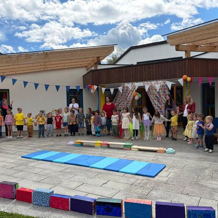 Eine Gruppe von Kindern, in bunten Kleidern gekleidet, versammelt sich vor einem Gebäude, einige halten Händchen. Bunte Wimpel und Luftballons schmücken den Bereich. Auf dem Boden liegen Matten, Blöcke und eine Bank. Erwachsene sind anwesend und beaufsichtigen.