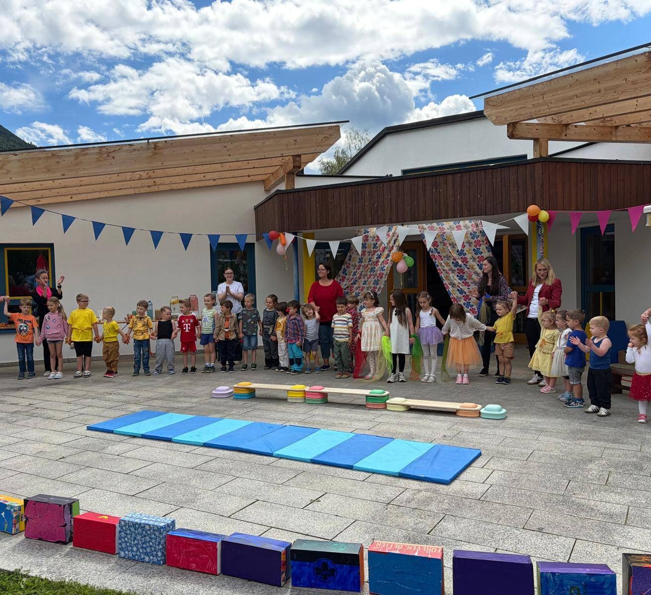 Eine Gruppe von Kindern, in bunten Kleidern gekleidet, versammelt sich vor einem Gebäude, einige halten Händchen. Bunte Wimpel und Luftballons schmücken den Bereich. Auf dem Boden liegen Matten, Blöcke und eine Bank. Erwachsene sind anwesend und beaufsichtigen.