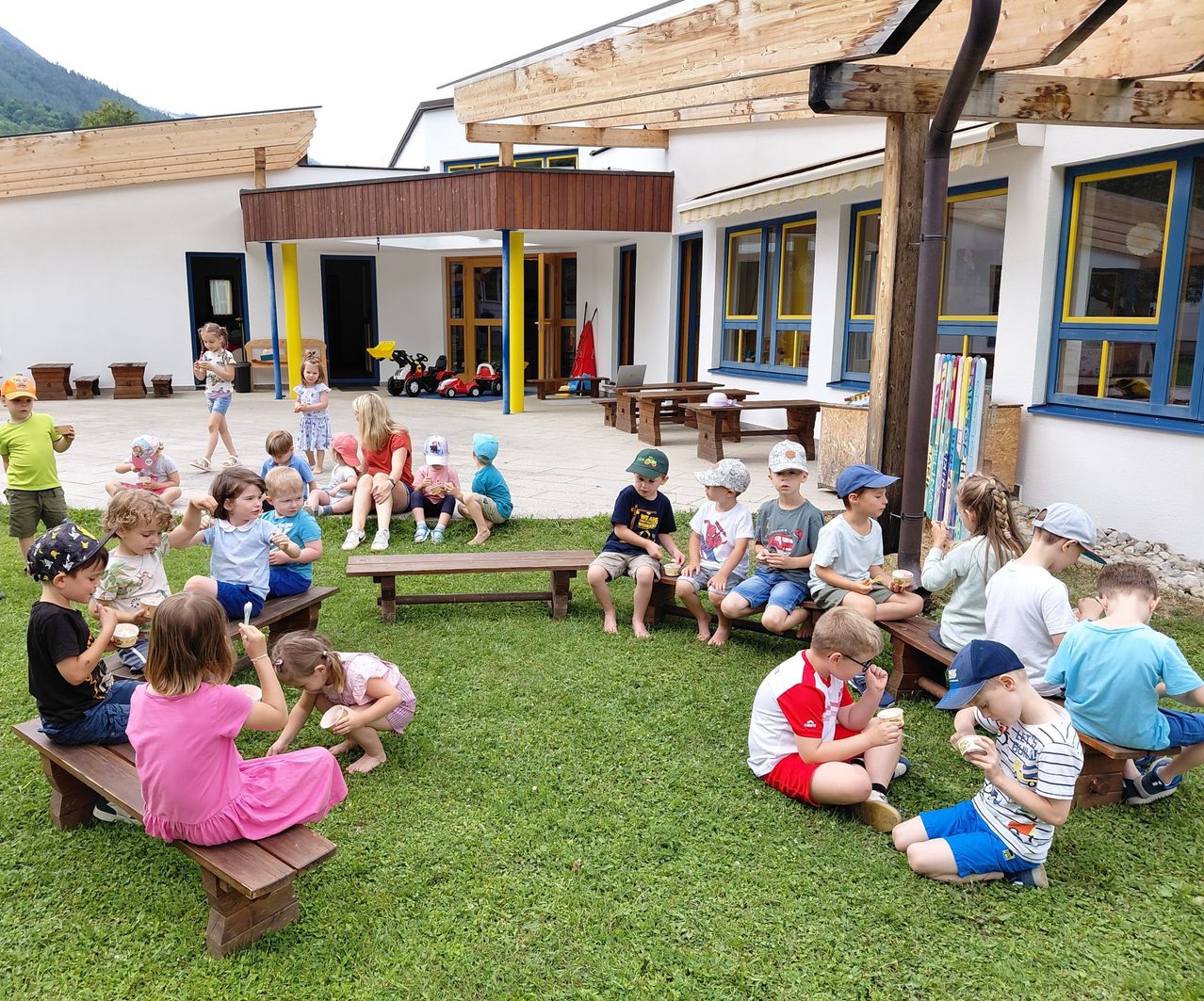 Eine Gruppe von Kindern und Erwachsenen sitzt auf Bänken im Hof. Sie essen Eis. Ein Gebäude mit Glasfenstern steht hinter ihnen.