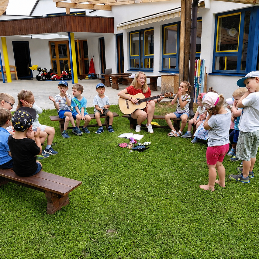 Eine Frau spielt eine Gitarre vor Kindern, die auf Bänken in einer Rasenfläche sitzen. Die Kinder hören aufmerksam zu, und es liegen Spielzeug und Gegenstände auf dem Boden.