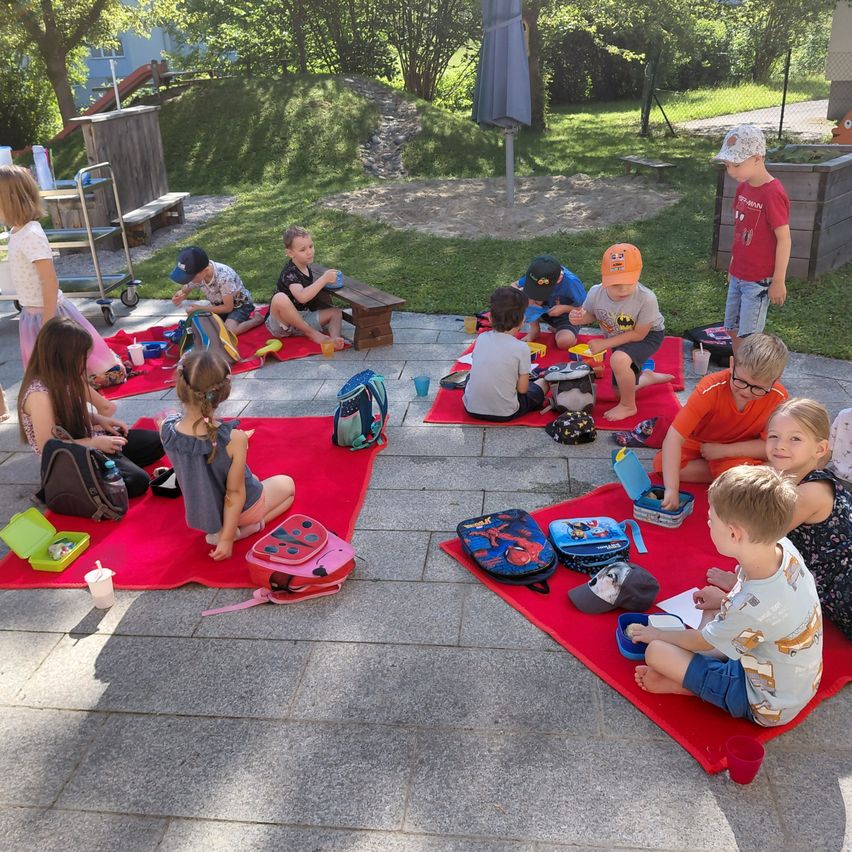 Kinder sitzen auf roten Decken im Freien. Einige essen und andere haben ihre Schulranzen in der Nähe. Ein Junge steht im Hintergrund in der Nähe einer Bank.