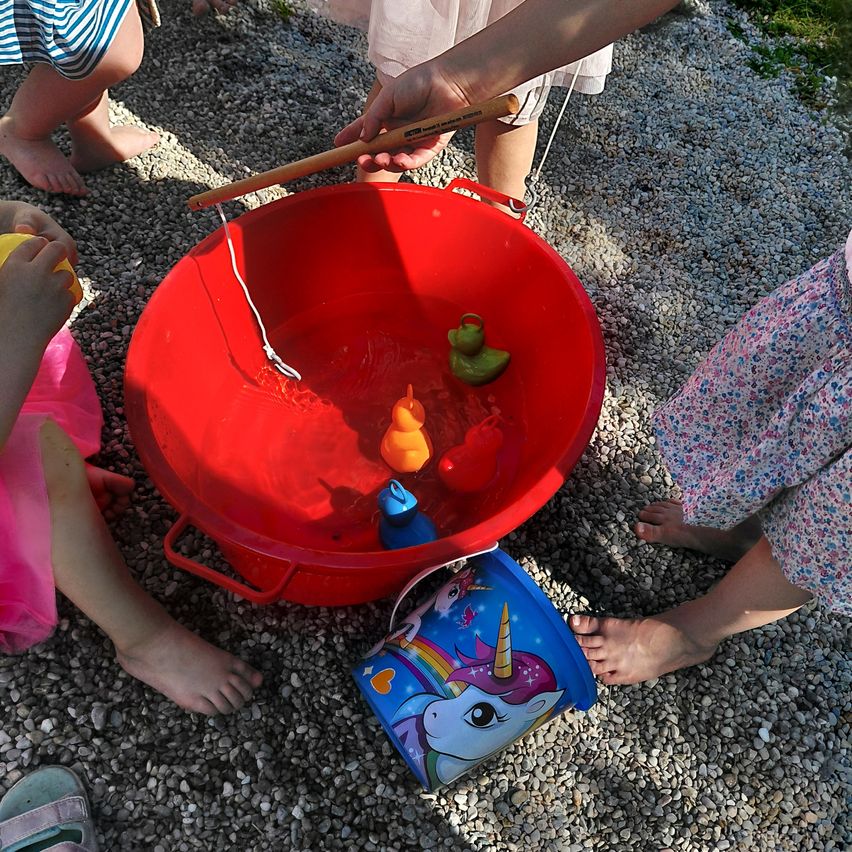 Eine Gruppe von Kindern spielt in einem roten Eimer mit Wasser. Sie benutzen einen Stock, um Spielzeug darin zu fangen. Ein Einhorn-Eimer steht in der Nähe.