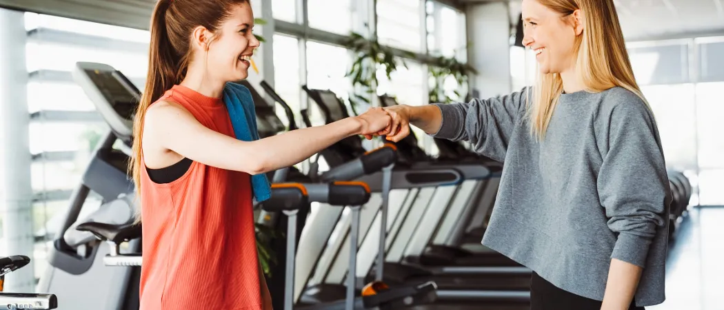 Zwei Frauen schütteln sich in einem Fitnessstudio die Hände, eine trägt ein orangefarbenes Tanktop und die andere ein graues Langarmshirt. Sie lächeln und scheinen Freundinnen zu sein.