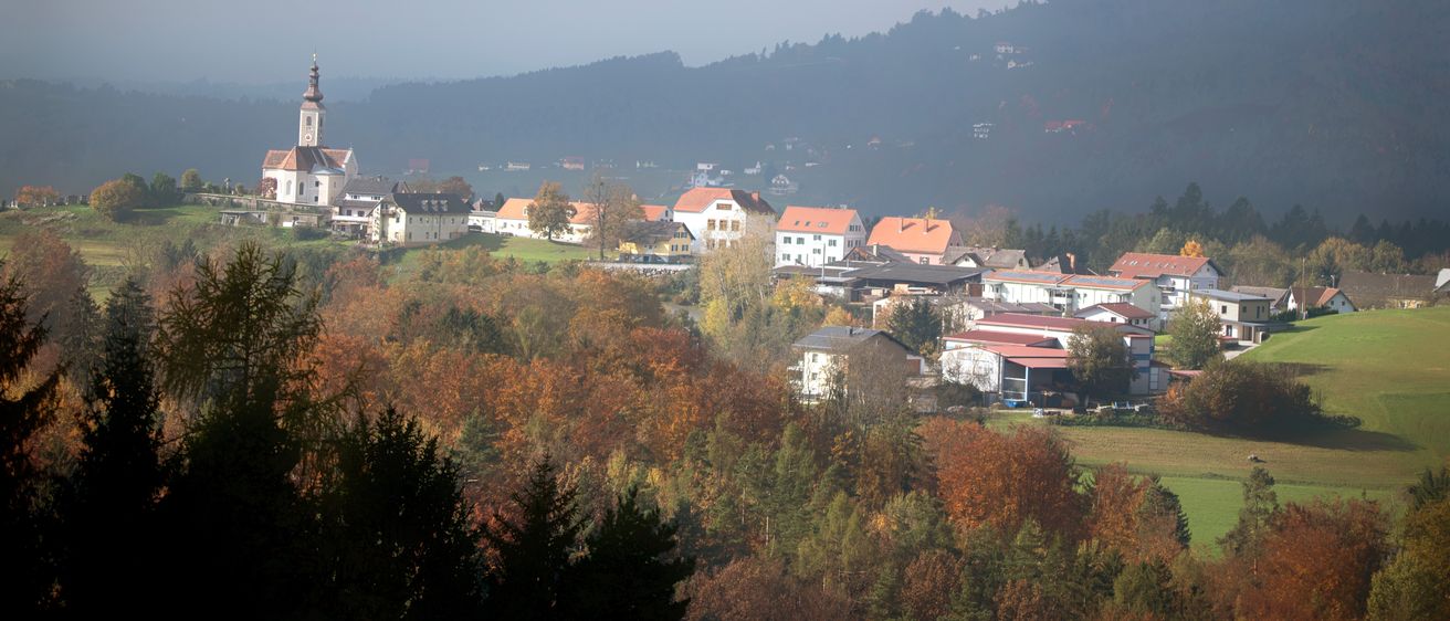 Ein nebeliger Morgenblick auf ein kleines Dorf, eingebettet zwischen Bäumen und Bergen, mit einem Kirchturm in der Ferne sichtbar.