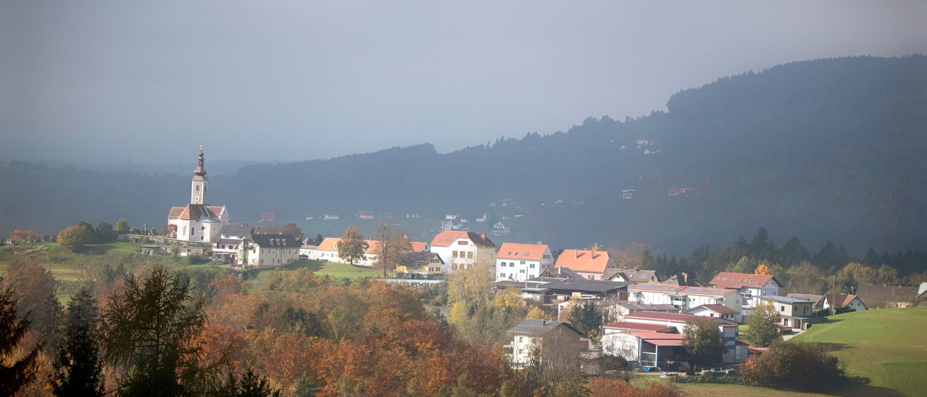 Ein nebeliger Morgenblick auf ein kleines Dorf, eingebettet zwischen Bäumen und Bergen, mit einem Kirchturm in der Ferne sichtbar.