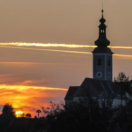 Eine Silhouette einer Kirche mit Turm und Uhren steht vor einem orange- und gelbtönten Sonnenuntergangshimmel mit Kondensstreifen am Himmel.