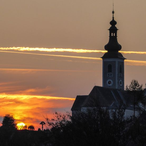 Eine Silhouette einer Kirche mit Turm und Uhren steht vor einem orange- und gelbtönten Sonnenuntergangshimmel mit Kondensstreifen am Himmel.