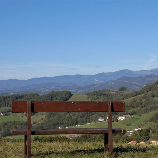 Eine Holzbank befindet sich auf einem grasbewachsenen Hügel mit Panoramablick auf die Berge und Täler. Der Himmel ist klar und blau.