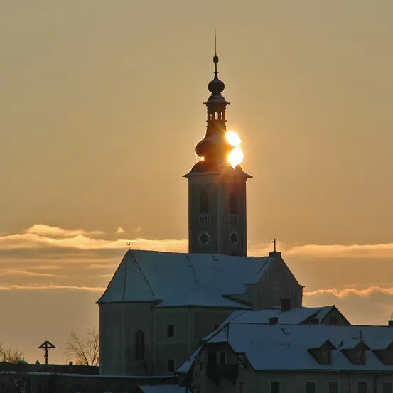 Die Sonne geht hinter einem Kirchturm unter und erhellt ihn, während sie ein warmes Licht auf die verschneiten Dächer wirft.