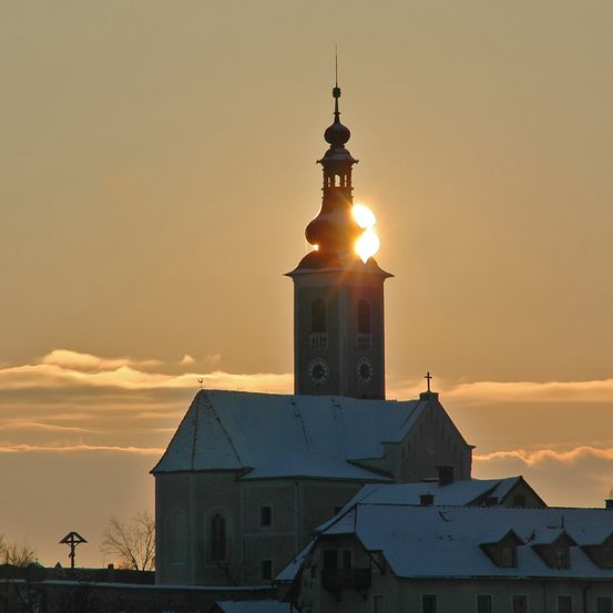 Die Sonne geht hinter einem Kirchturm unter und erhellt ihn, während sie ein warmes Licht auf die verschneiten Dächer wirft.