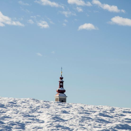 Ein Kirchturm erhebt sich auf einem verschneiten Hügel unter einem blauen Himmel mit verstreuten Wolken.