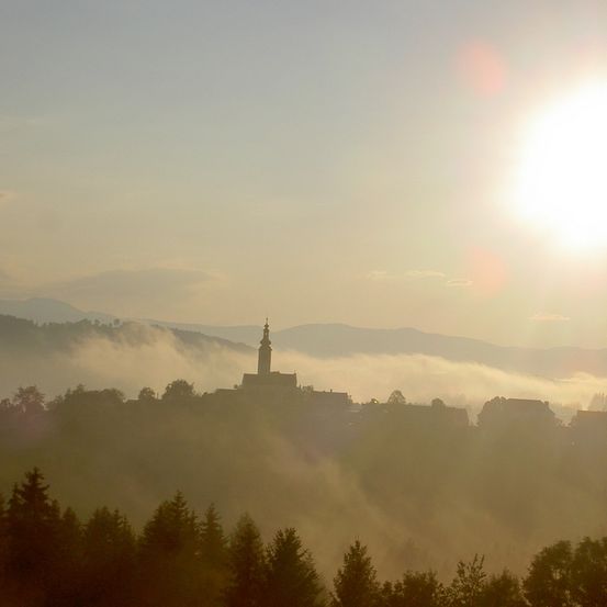 Ein nebliger Morgen mit aufgehender Sonne über einer Stadt auf einem Hügel mit einem Turm in der Mitte, umgeben von Bäumen und Bergen.