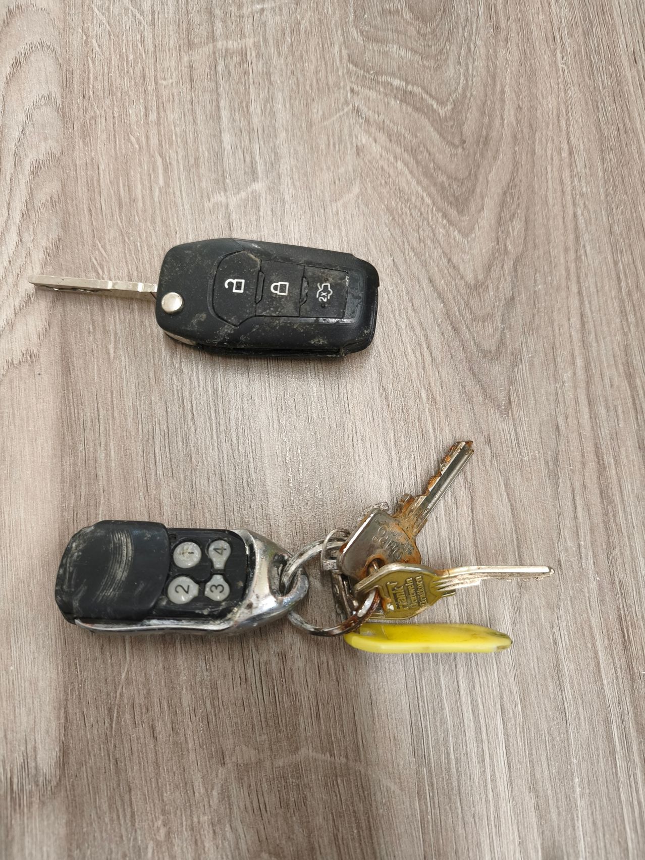 Two car keys are placed on a wooden surface. The top key has a silver blade and a black remote control with buttons. The bottom key has a silver blade and a black remote control with a keychain.