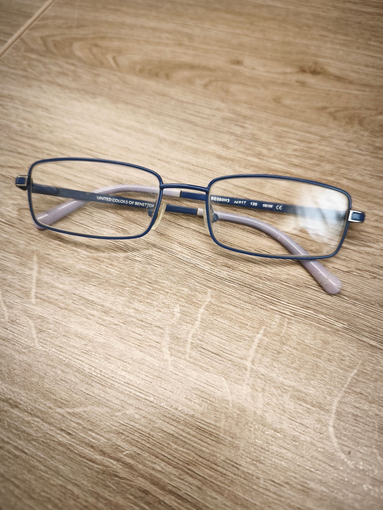 A pair of blue-framed glasses with clear lenses rests on a wooden surface. The brand name 'UNITED COLORS OF BENETTON' is inscribed on the left lens.