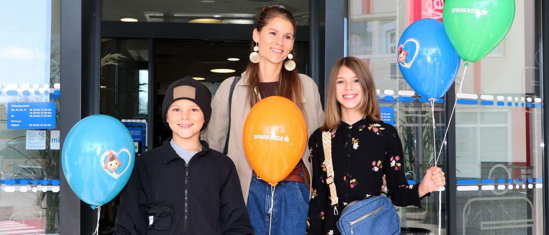 Drei Personen, darunter eine Frau und zwei Kinder, stehen lächelnd vor einem Gebäude und halten Luftballons. Die Frau trägt Ohrringe und einen beigefarbenen Mantel. Die Kinder halten orangefarbene und blaue Luftballons. Hinter ihnen befindet sich eine Glastür mit einem Mülleimer.