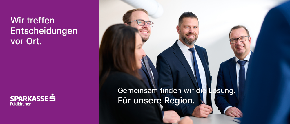 Vier Geschäftsleute in formeller Kleidung lächeln und stehen um einen Schreibtisch herum, auf dem ein Stift liegt. Eine Frau schaut einen Mann an, der schreibt. Der Text lautet: 'Gemeinsam finden wir die Lösung. Für unsere Region.'