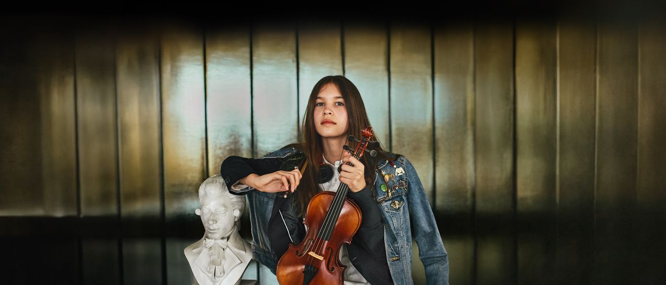 Ein junges Mädchen in einer Jeansjacke posiert mit einer Geige vor einer Büste von Beethoven.