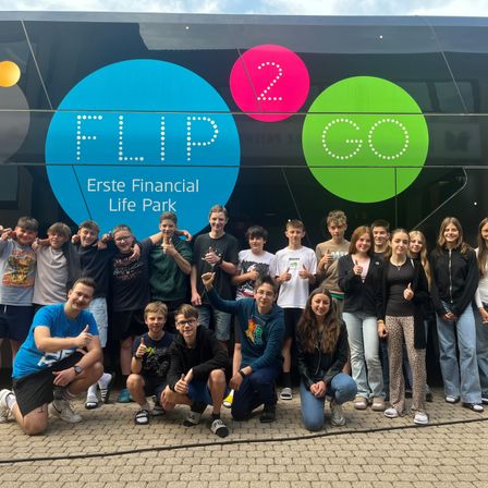 Eine Gruppe von Menschen posiert für ein Foto vor einem Bus mit den Worten 'Erste Financial Life Park' und den Zahlen '2 Go'. Sie lächeln und geben Daumen hoch.