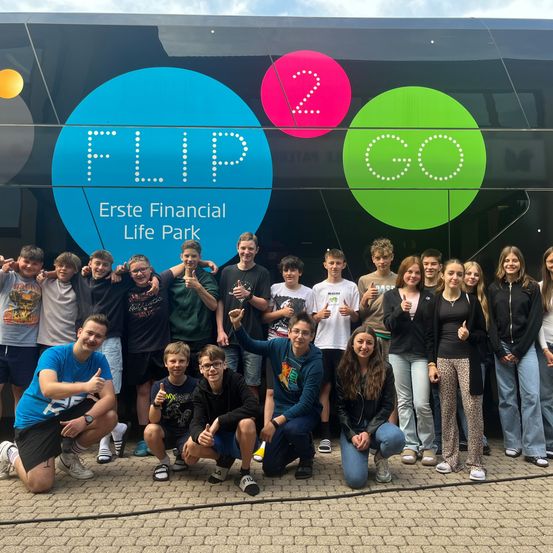 Eine Gruppe von Menschen posiert für ein Foto vor einem Bus mit den Worten 'Erste Financial Life Park' und den Zahlen '2 Go'. Sie lächeln und geben Daumen hoch.