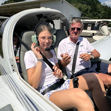 Ein junges Mädchen mit Brille und Kopfhörern sitzt in einem kleinen Flugzeug, lächelt, während ein älterer Mann neben ihr eine Sonnenbrille und eine Uhr trägt.