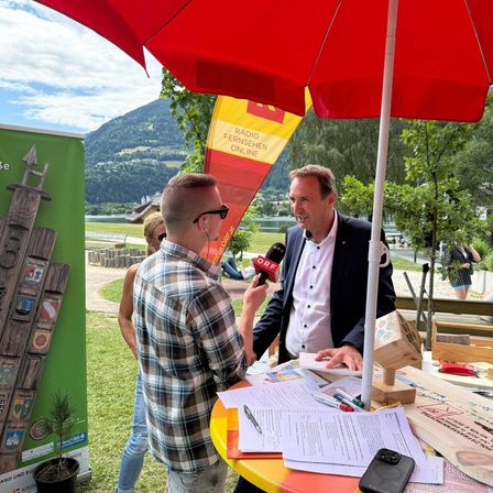 Zwei Männer stehen im Freien unter einem roten Schirm. Ein Mann hält ein Mikrofon und interviewt wahrscheinlich den anderen Mann im Anzug. Sie sitzen an einem Tisch mit Papieren, einem Handy und Stiften. Ein Berg und ein See sind im Hintergrund.