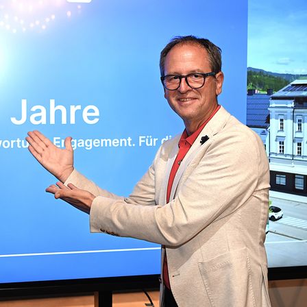 Ein lächelnder Mann mit Brille und heller Jacke steht vor einem großen Bildschirm und gestikuliert mit den Händen. Der Bildschirm hinter ihm zeigt eine Nachricht in Deutsch.