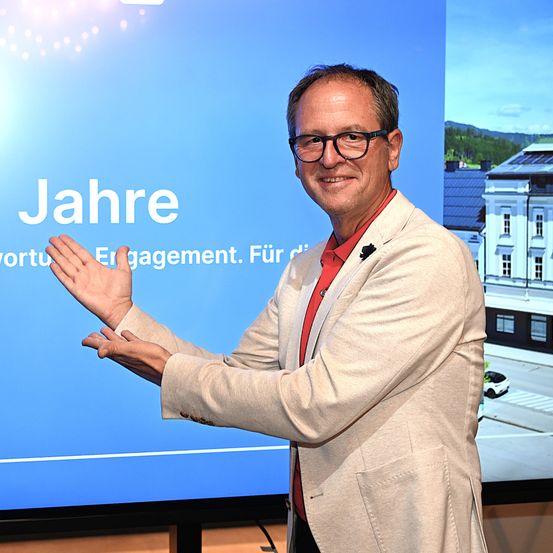 Ein lächelnder Mann mit Brille und heller Jacke steht vor einem großen Bildschirm und gestikuliert mit den Händen. Der Bildschirm hinter ihm zeigt eine Nachricht in Deutsch.