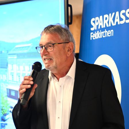 Ein Mann in einem Anzug spricht in ein Mikrofon vor einer großen Leinwand, die ein Gebäude und die Worte 'Sparkasse Feldkirchen' zeigt.