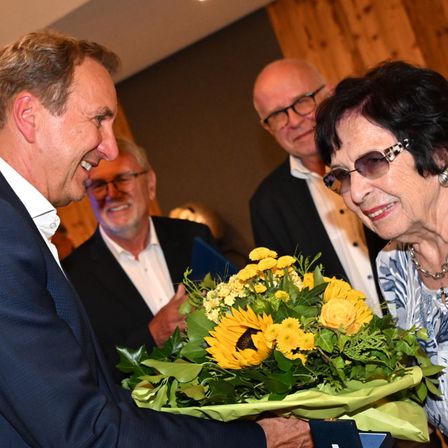 Ein Mann in einem Anzug reicht einer älteren Frau einen Strauß Sonnenblumen, während zwei andere Männer in Anzügen zusehen.