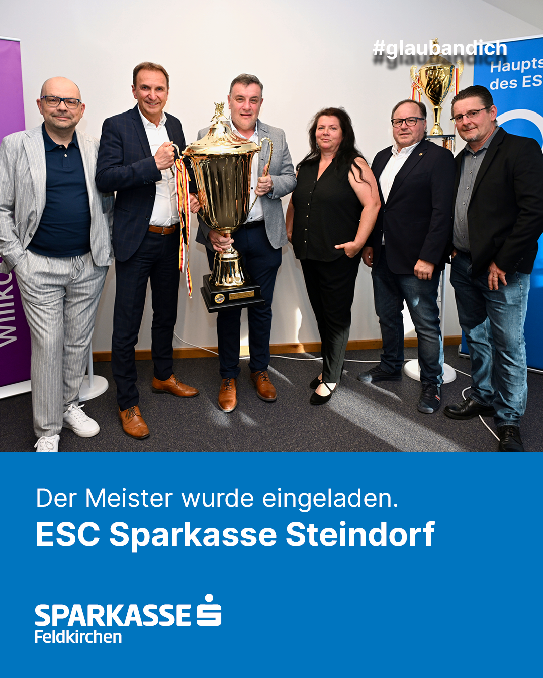 Eine Gruppe von Menschen steht zusammen und hält eine Trophäe, mit deutschem Text 'Der Meister wurde eingeladen.' Sie alle scheinen zu lächeln.