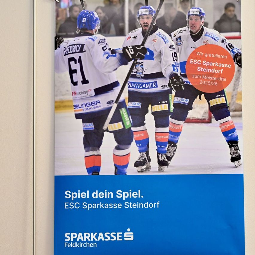 Plakat von Eishockey-Spielern mit Trikots mit den Nummern 61, 19 und 26. Sie feiern auf der Eisbahn mit einem roten kreisförmigen Etikett, das 'Wir gratulieren ESC Sparkasse Steindorf zum Meistertitel 2025/26' sagt. Unten steht 'Spiel dein Spiel. ESC Sparkasse Steindorf'.