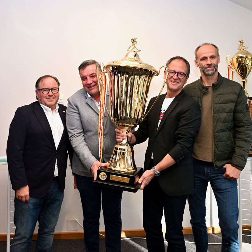 Fünf Männer stehen lächelnd in einem Raum, einer hält eine Trophäe. Sie sind in Anzügen und mit Brillen gekleidet. Hinter ihnen sind zwei Trophäen ausgestellt.