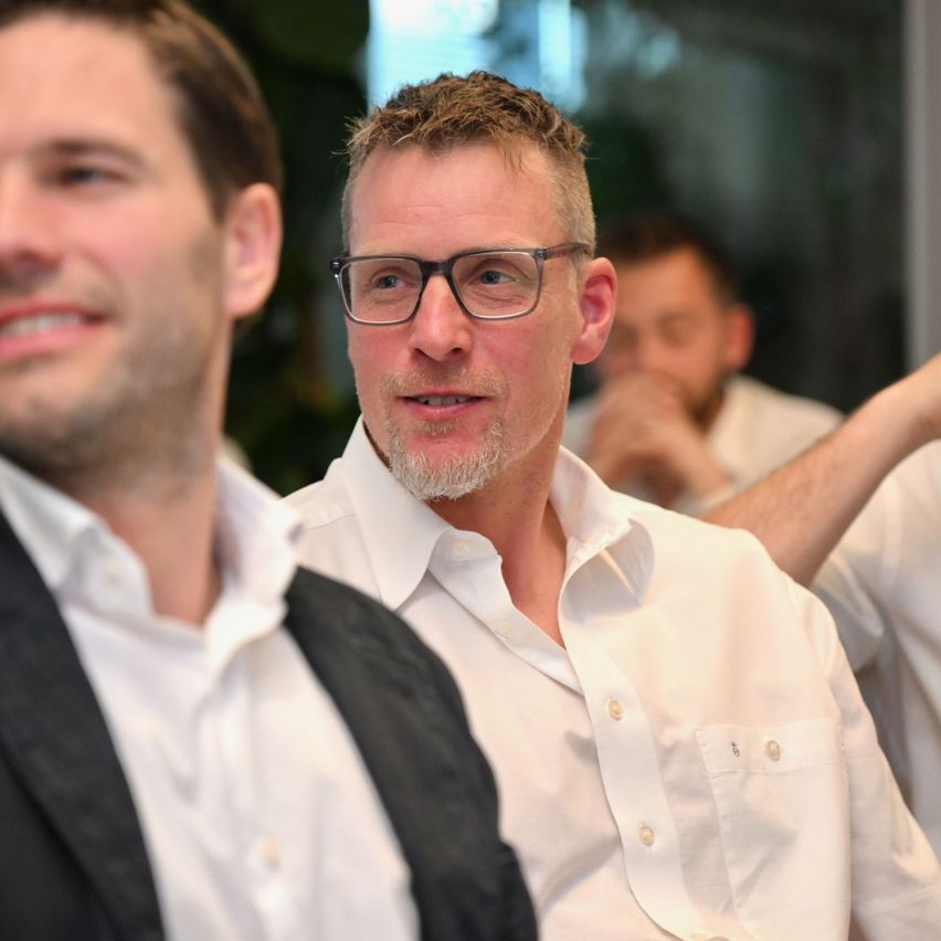 Zwei Männer sind drinnen, möglicherweise bei einer Besprechung. Der Mann mit Brille spricht, während der andere Mann aufmerksam zuhört. Dahinter ist ein weiterer Mann verschwommen und scheint Notizen zu machen.