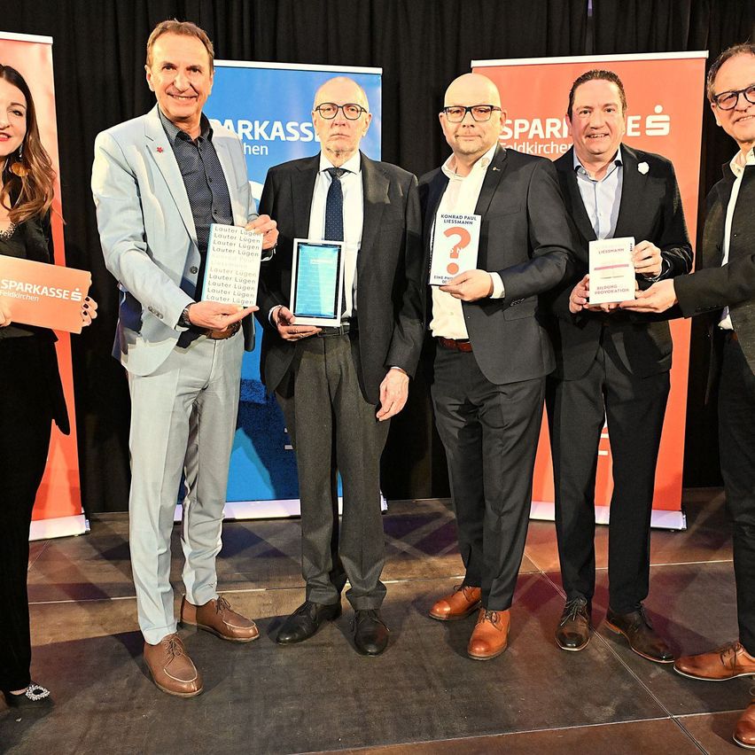 Fünf Männer und eine Frau stehen lächelnd zusammen auf einer Bühne. Jeder hält ein Buch. Der Mann ganz links hält ein Buch mit dem Titel 'Lautke Eugen'. Die Frau hält ein Buch mit dem Titel 'Sparkasse'.
