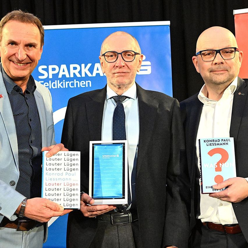 Drei Männer in Anzügen stehen nebeneinander vor einem Banner mit der Aufschrift 'SPARKASSE Feldkirchen'. Der Mann auf der linken Seite hält ein Buch mit dem Titel 'Lauter Lügen'. Der Mann in der Mitte hält ein Tablet mit blauer Abdeckung. Der Mann auf der rechten Seite hält ein Buch mit dem Titel 'Was Nun?'.