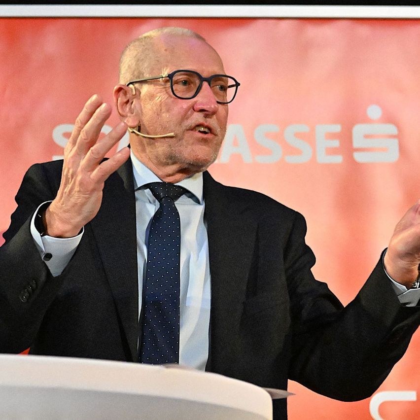 Ein älterer Mann in einem schwarzen Anzug und Krawatte hält eine Rede, gestikuliert mit den Händen und trägt eine Brille und ein Mikrofon.