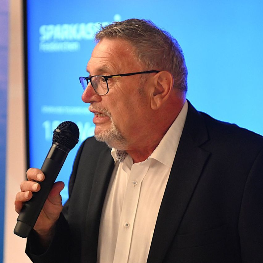 Ein Mann in einem schwarzen Anzug und mit Brille spricht in ein Mikrofon bei einer Veranstaltung.