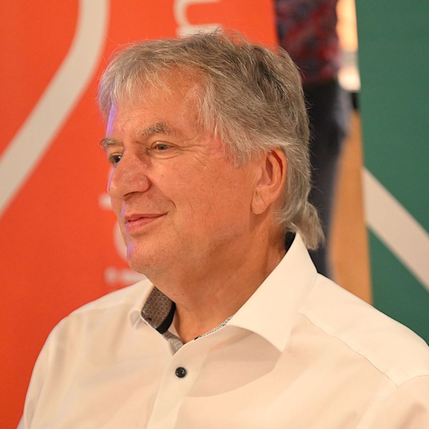 Ein Mann mit grauem Haar und einem weißen Hemd lächelt in die Kamera und steht vor einem unscharfen Hintergrund mit einem orangefarbenen und grünen Banner.