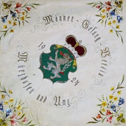 Ein antikes Taschentuch zeigt in der Mitte ein Wappen, umgeben von einer Blumenbordüre. Der Text lautet 'Manne-Belang 19 24' und 'Wilhelmen und Unf'. Das Wappen ist grün mit einem weißen Vogel.