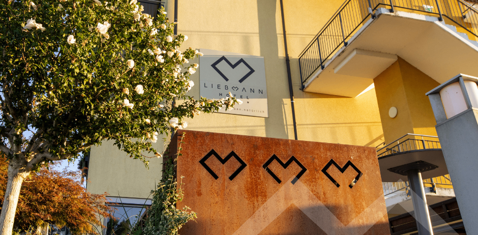 Die Vorderseite des Liebmann Hotels in Gelb mit einem Balkon und einem Baum davor. Ein Schild mit den Buchstaben MMM steht vor dem Gebäude.