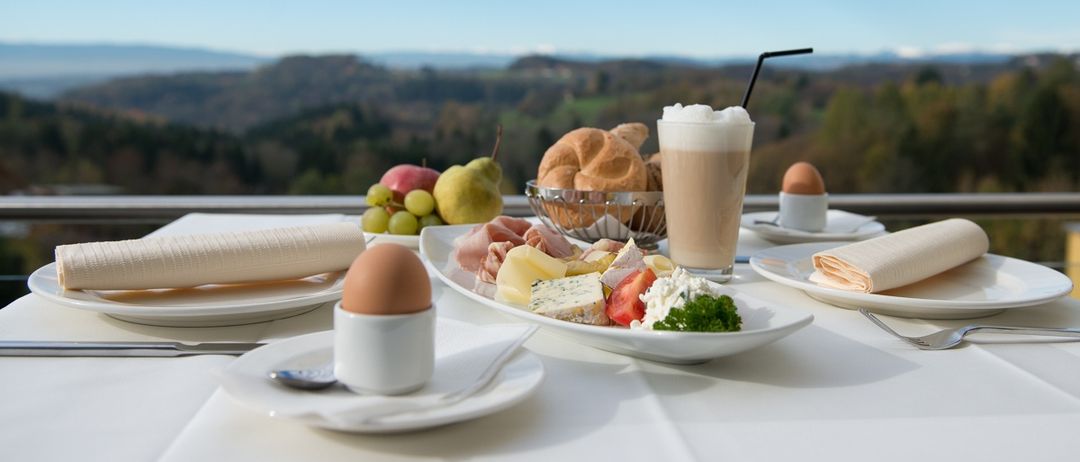 Ein Frühstückstisch mit Blick auf Berge, bestehend aus Eiern, Brot, Käse, Obst und einem Kaffeegetränk.