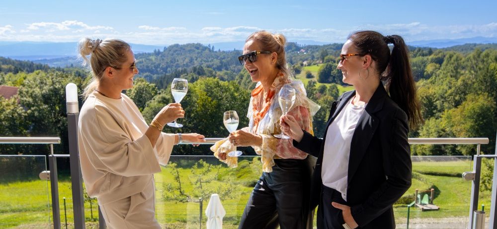 Drei Frauen auf einem Balkon mit Blick auf eine üppige, grüne Landschaft stoßen mit Weinläsern an und lächeln in die Kamera.